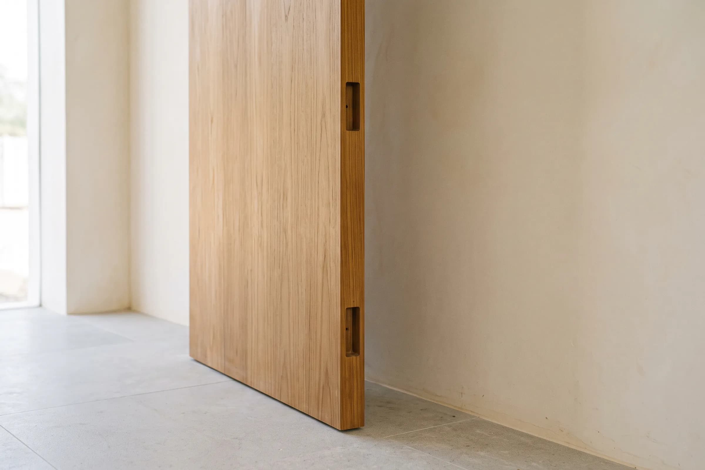 A finished door leaf standing on edge against a plaster wall, hinge edge to camera, showing its edge band and two empty hinge mortises.