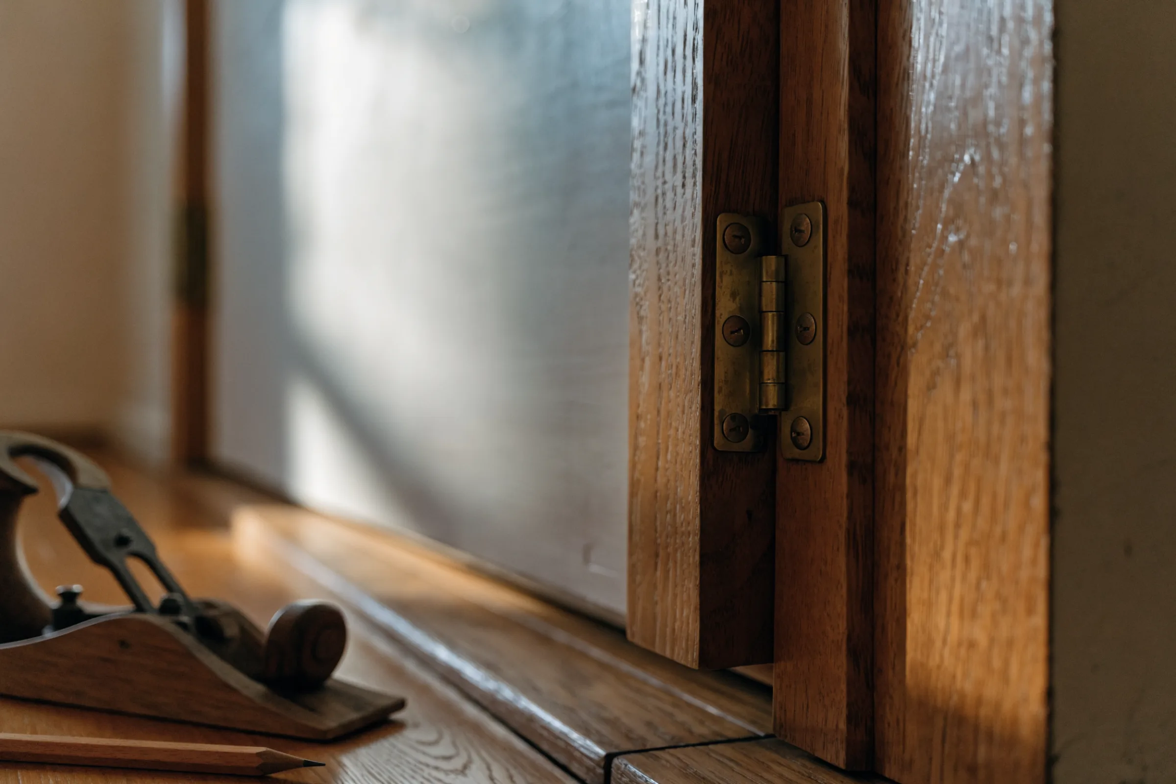 Close view of a brass butt hinge on a wooden frame edge beside obscured glass, with a hand plane and a pencil resting on the ledge below