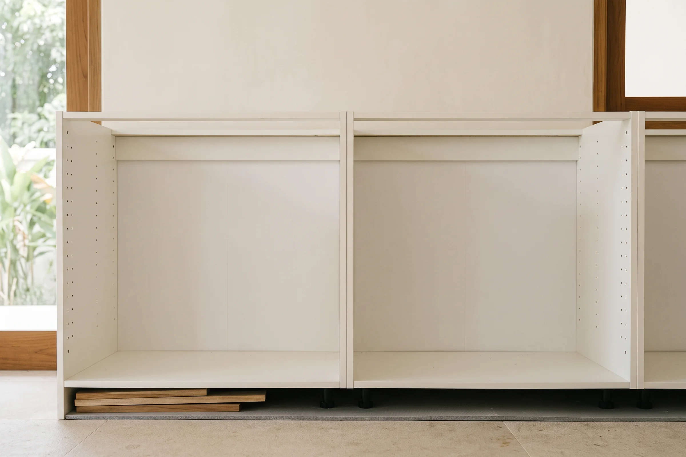 Two white flat-pack base carcasses standing against a wall without fronts, timber shims packed under one and adjustable feet under the other.