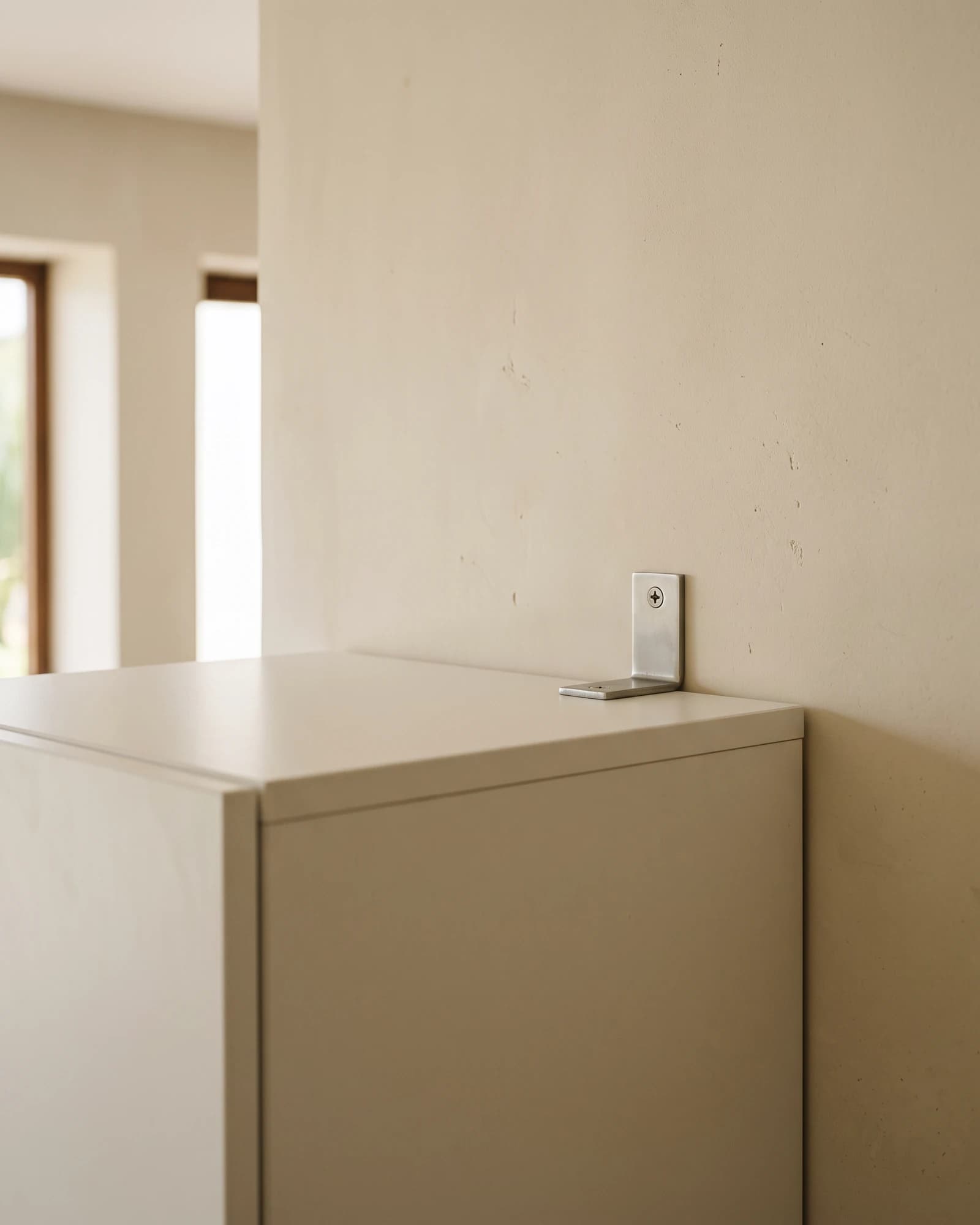 A small steel anti-tip bracket screwed to the top of a white wardrobe and to the plastered masonry wall behind it.