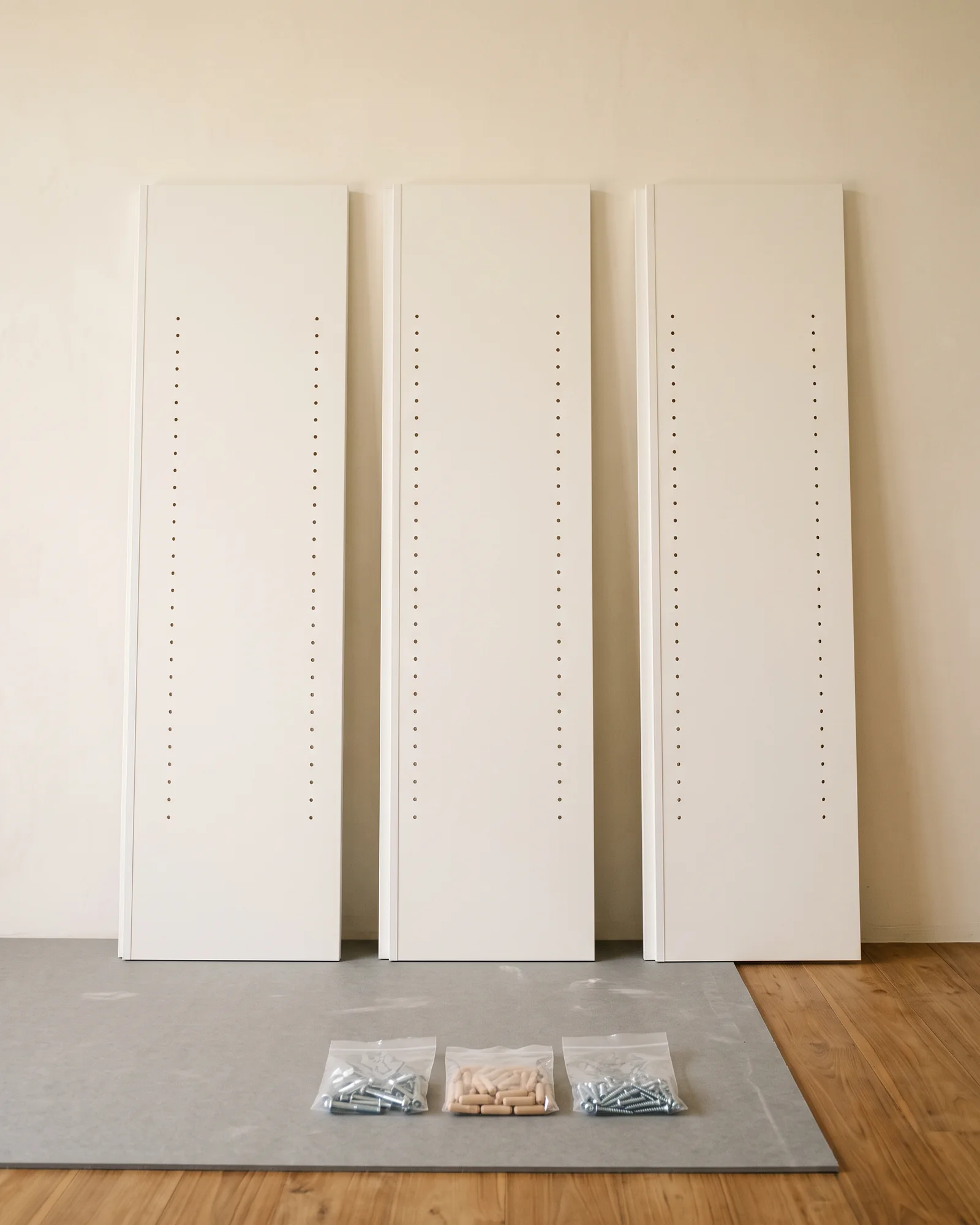 Three white flat-pack wardrobe panels standing upright against a wall, with clear bags of cam bolts, dowels and screws grouped on floor-protection board.
