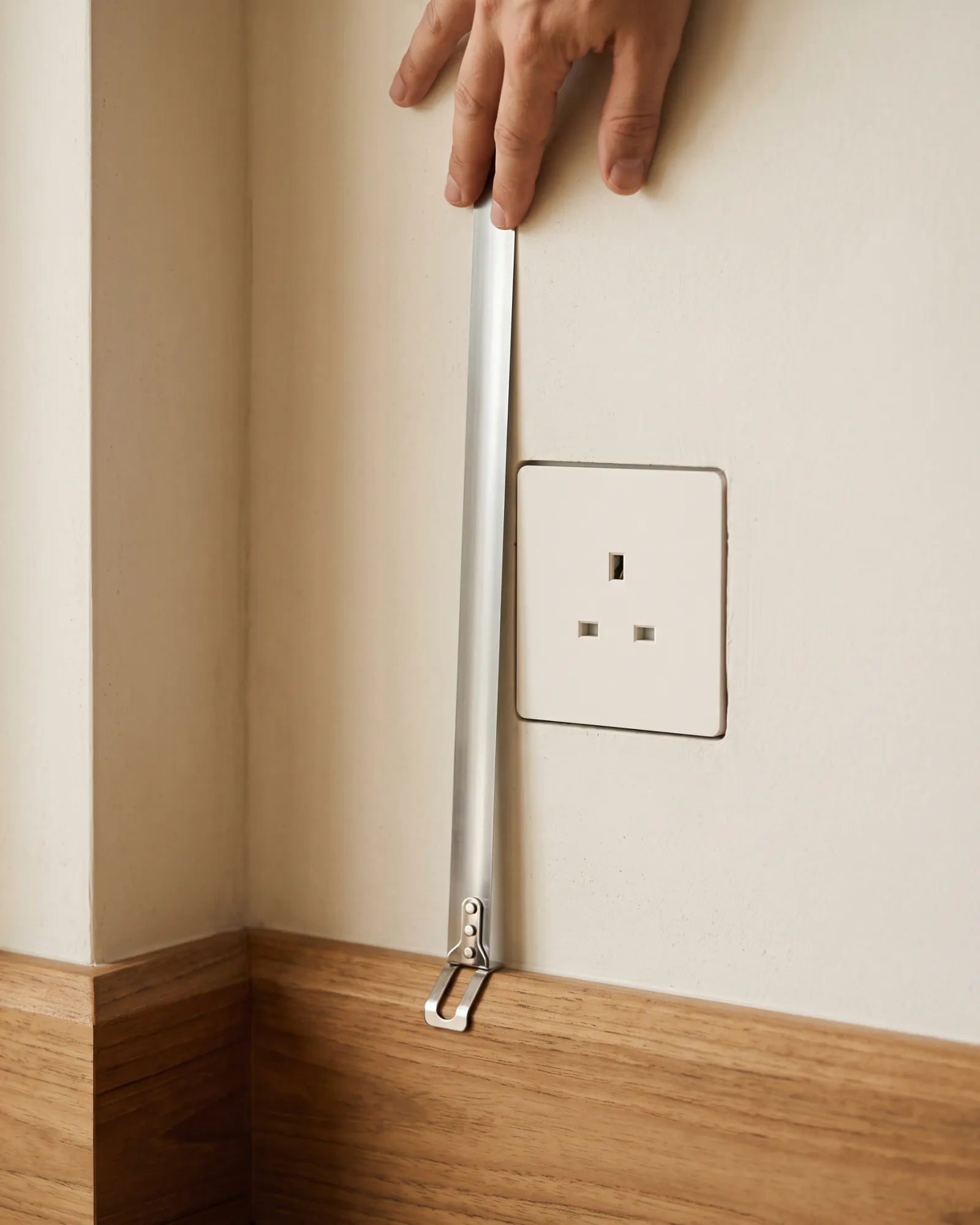 A tape measure held against a plaster wall in an alcove corner, its hook on a teak skirting board, a socket in the wall above.