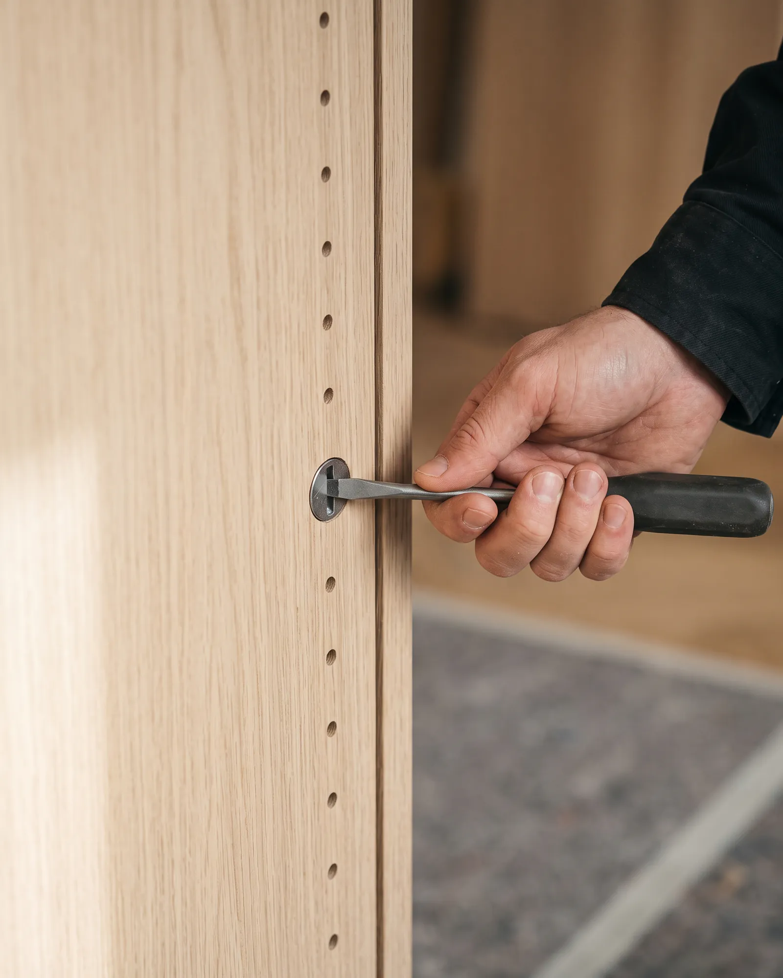 A hand turning a screwdriver in a cam lock set into a cabinet side panel, with the row of shelf-pin holes running down beside it.