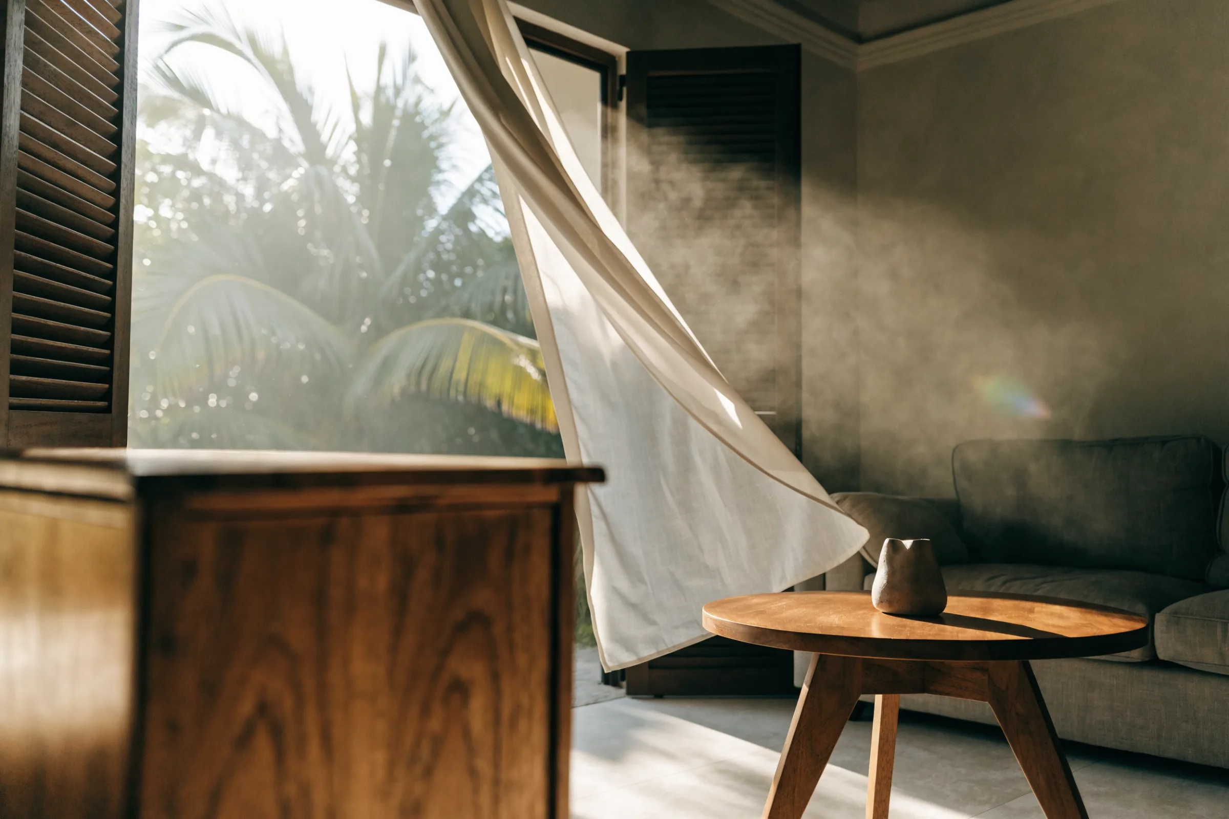 Teak sideboard beside an open window with a billowing white curtain and palm leaves outside
