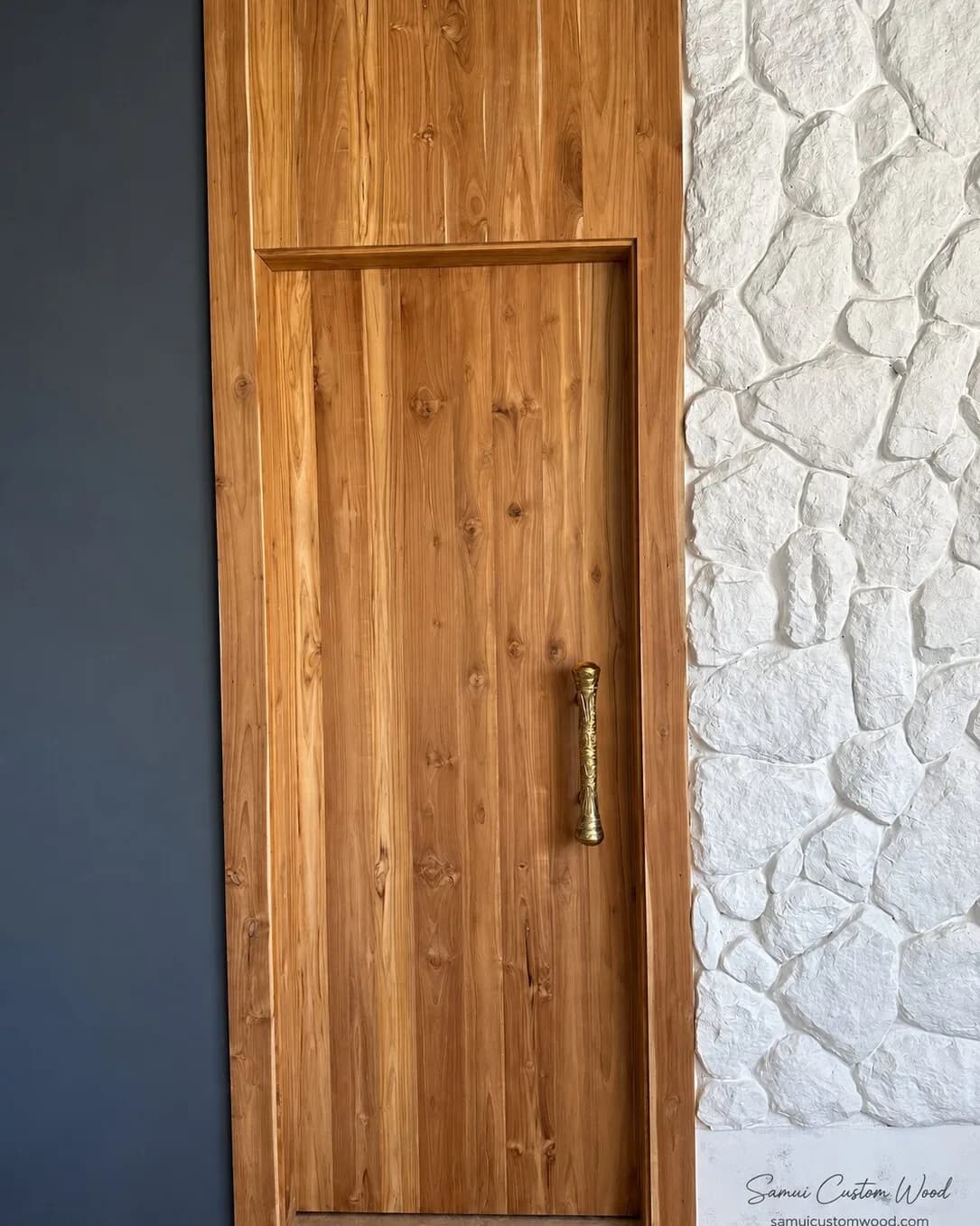 Teak plank door with a brass handle set between a stone-clad wall and dark panelling