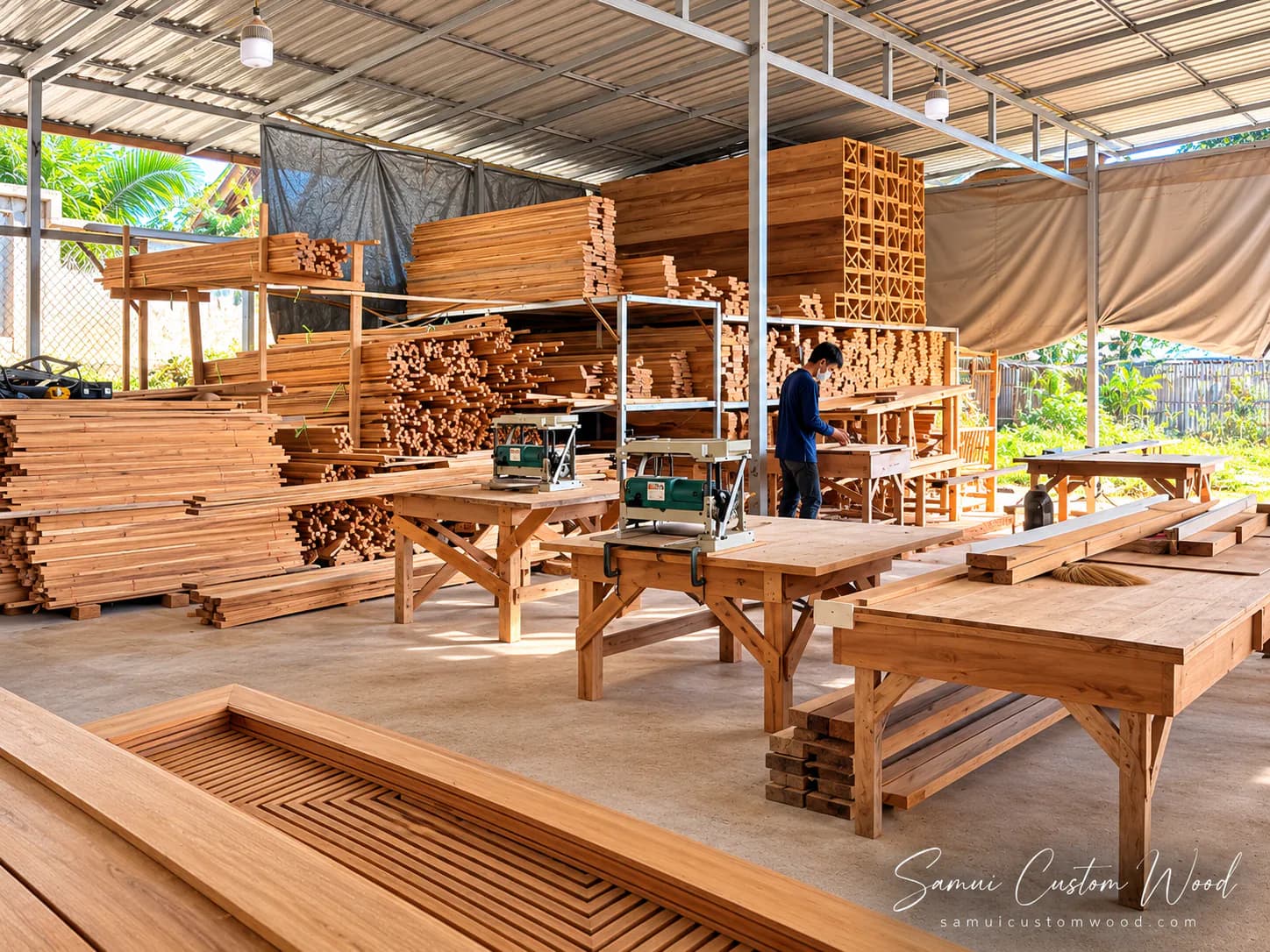 ภายในโรงงาน โต๊ะช่างพร้อมเครื่องไสไม้ ชั้นวางไม้สักแปรรูป และช่างกำลังทำงานอยู่ที่โต๊ะด้านใน
