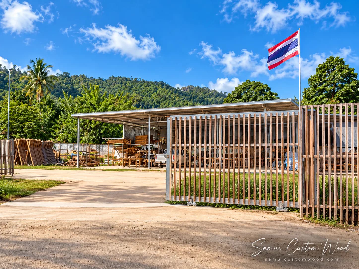 บริเวณโรงงานมองจากทางเข้า มีประตูรั้วไม้ระแนง โรงเปิดโล่งที่เก็บไม้และเฟอร์นิเจอร์ และเนินเขาอยู่ด้านหลัง